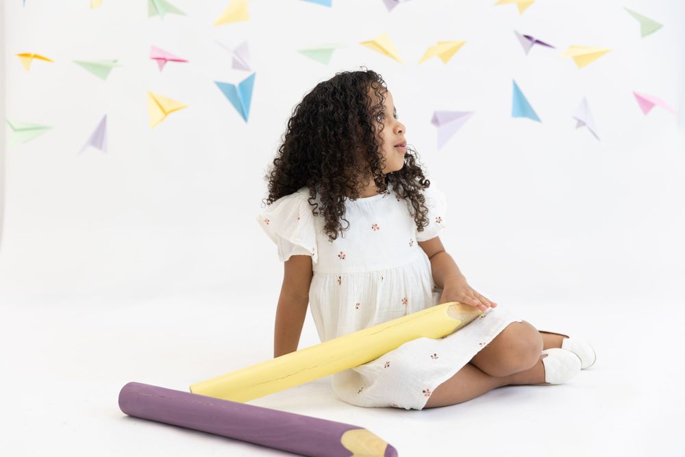 White-dressed child sitting on floor with large colorful pencils and confetti decorations against white backdrop.