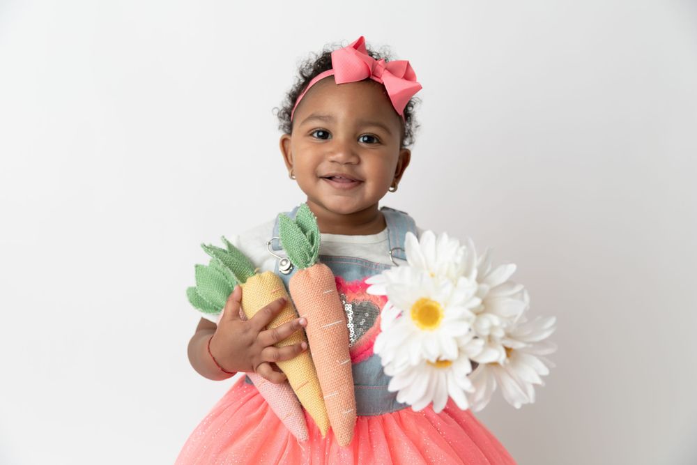 Child in tulle skirt holding decorative carrots and flowers against white background.