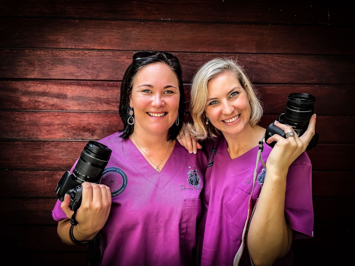 two birth photographers with cameras in purples scrubs in front of a wooden background blonde brunette