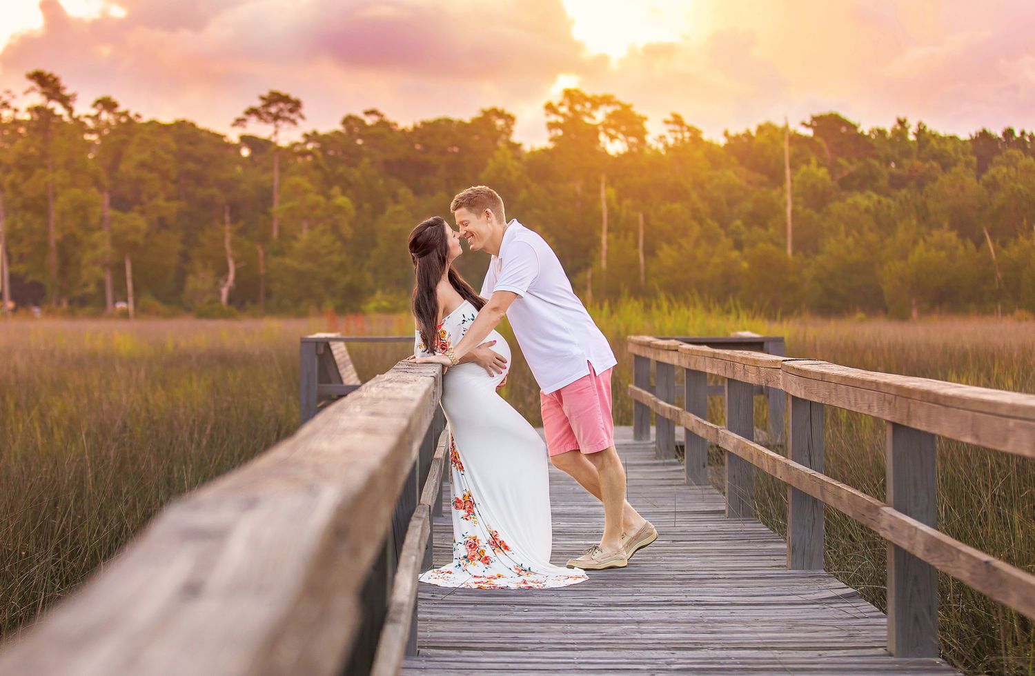 Couple embracing on wooden boardwalk during golden sunset with marsh landscape behind them.