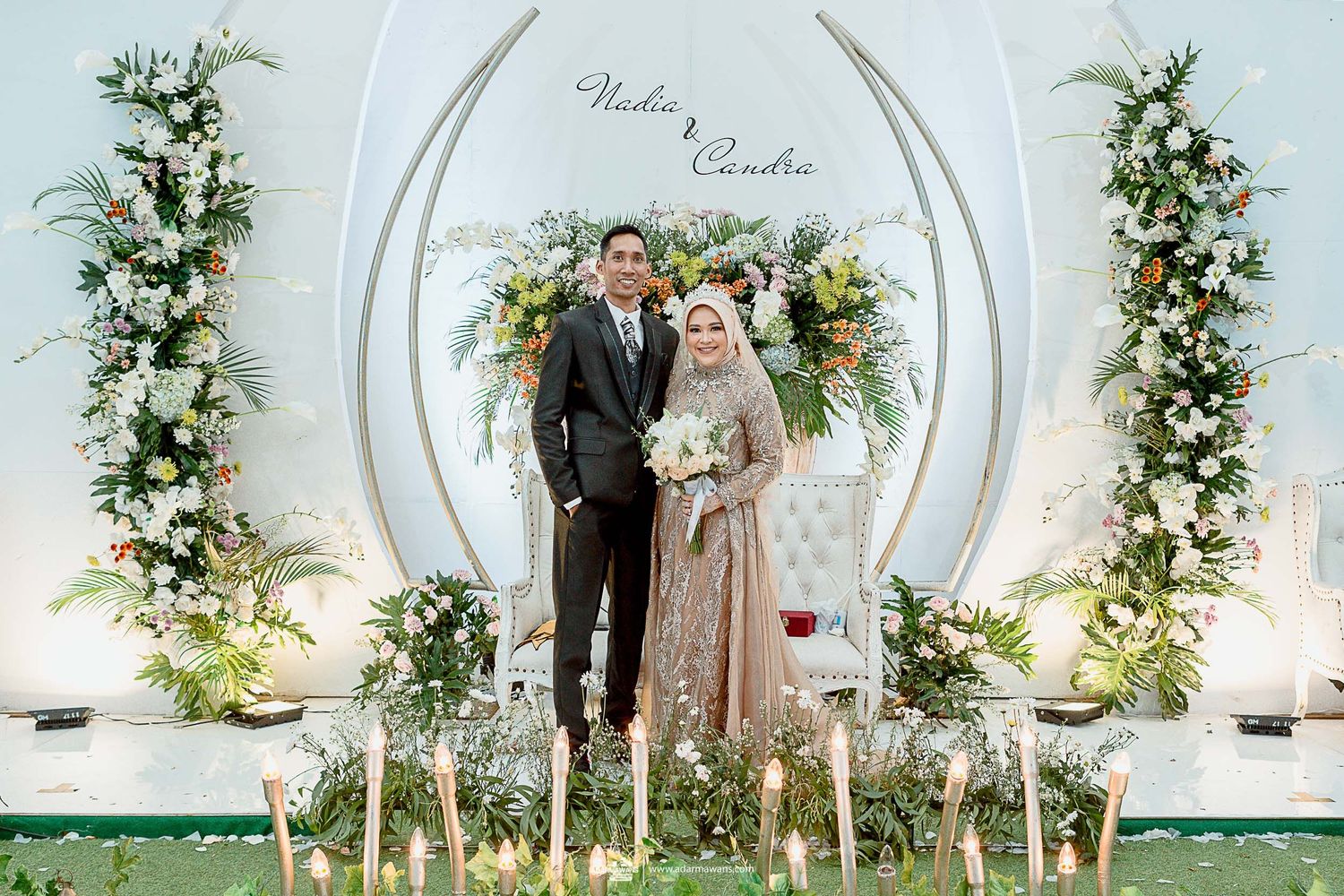 Wedding portrait in front of an elegant white arch decorated with fresh flowers and greenery.