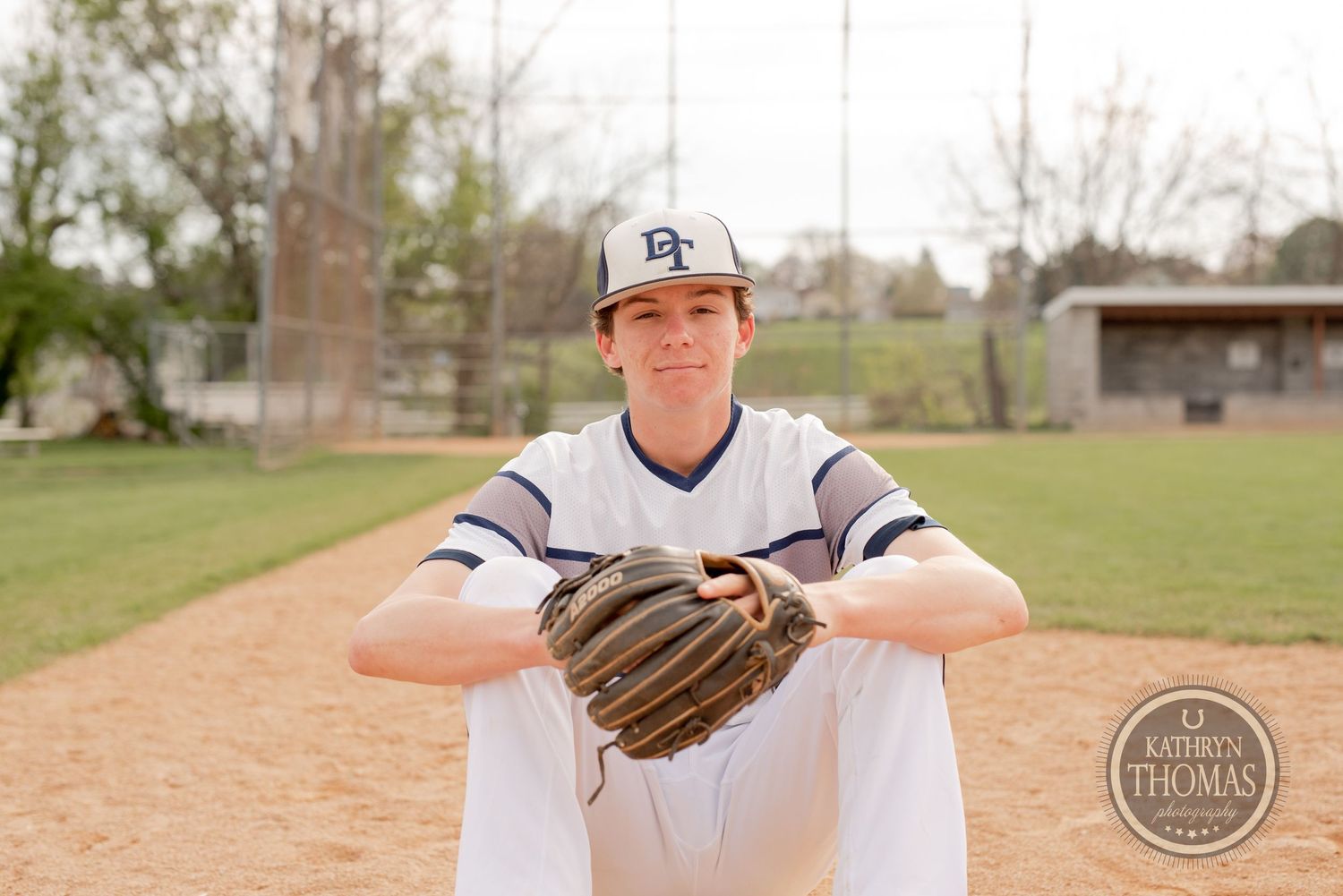Baseball player poses on dirt field with glove in white uniform with navy accents.