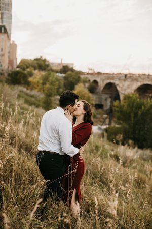 Couple sharing a passionate kiss in a field of tall grass with a historic stone arch bridge behind them.