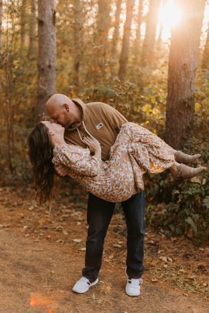 Man in tan hoodie romantically dipping and kissing woman in floral dress on a sunlit forest path in fall.