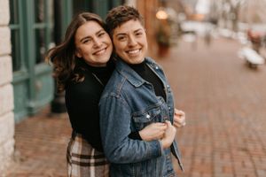 Two women smiling and embracing warmly on a brick sidewalk in a cozy urban neighborhood.