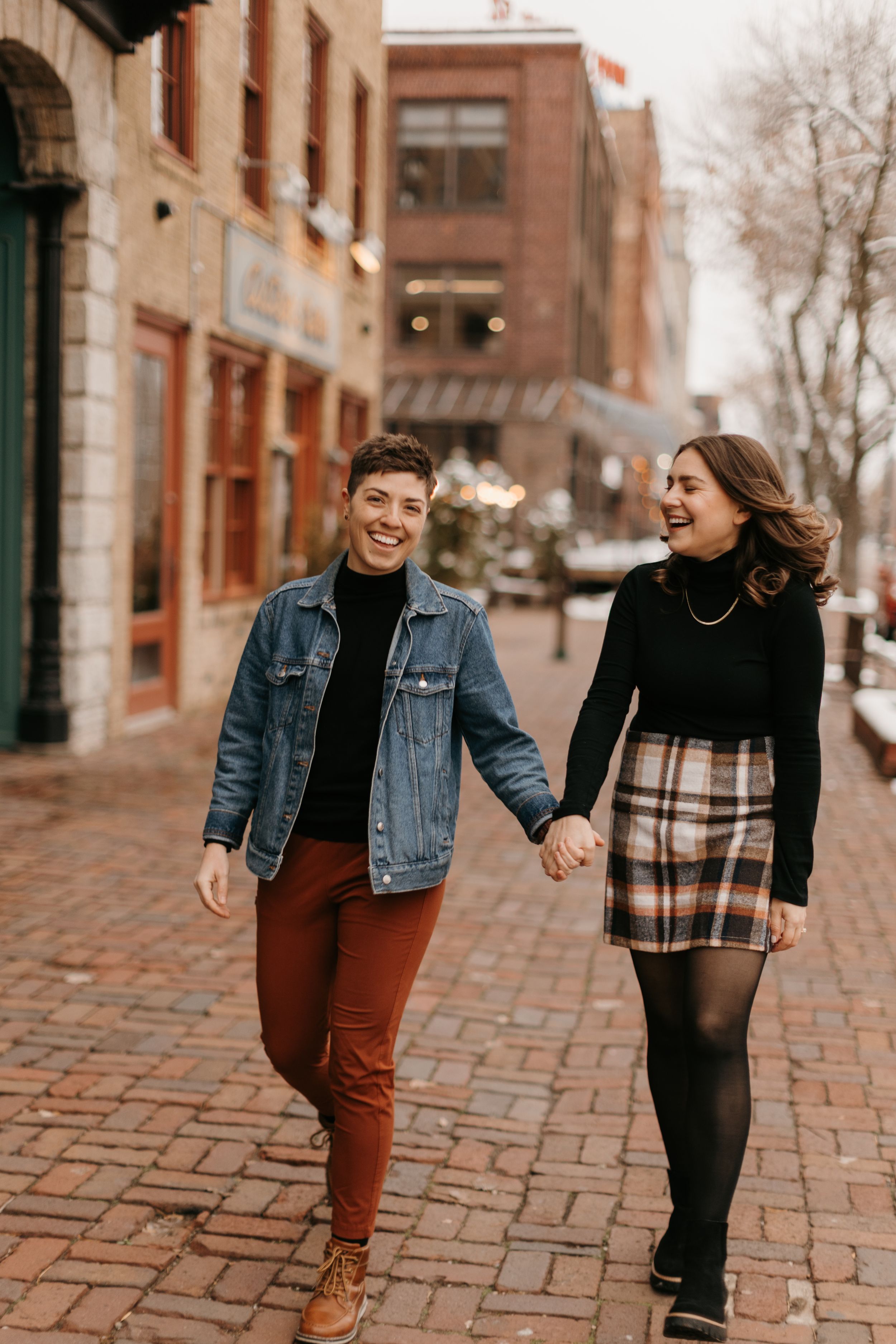 Happy couple holding hands and laughing while walking on a brick street in a charming urban setting.