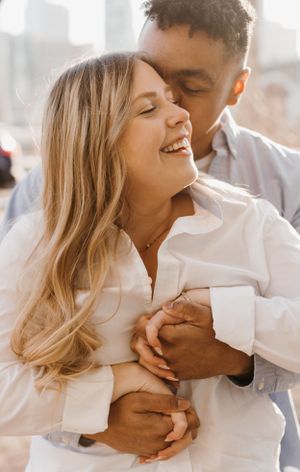 Man embracing woman from behind as she laughs joyfully during sunny outdoor engagement session.