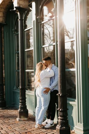 Couple sharing a kiss against elegant green storefront with sunlight glowing behind them.