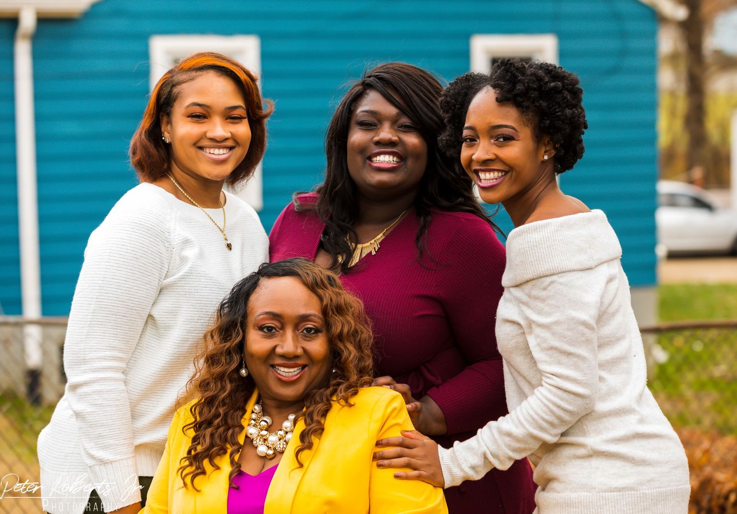 Four women smile together outdoors in front of a blue house, one wearing a bright yellow jacket.