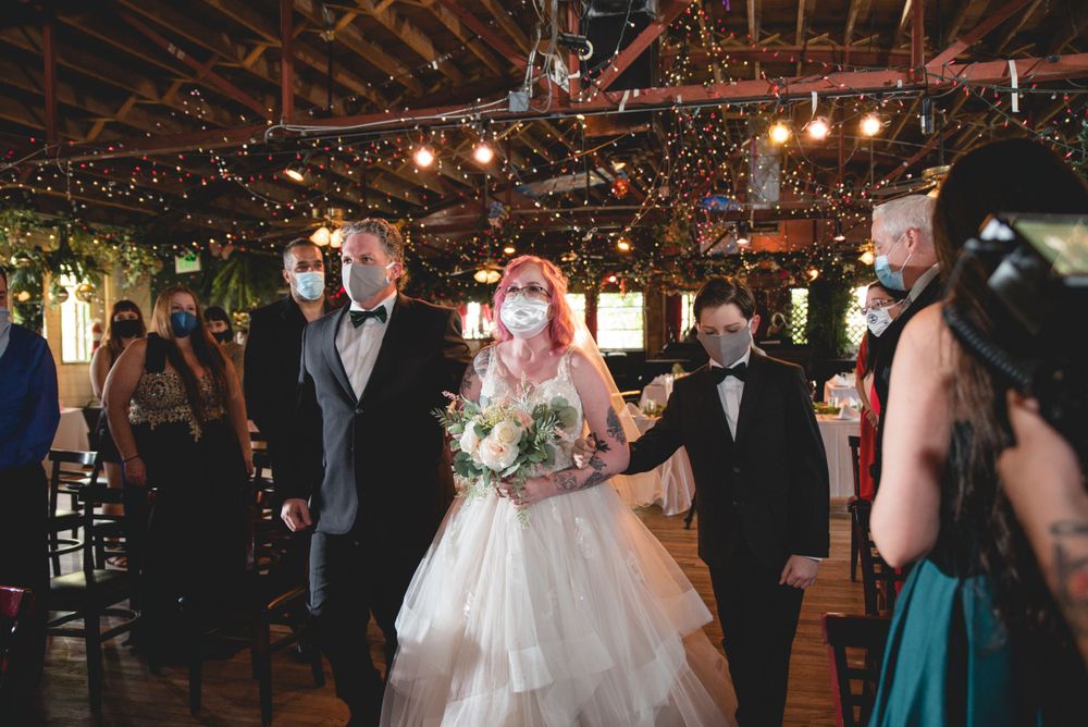 A wedding ceremony procession in a rustic venue with string lights overhead.