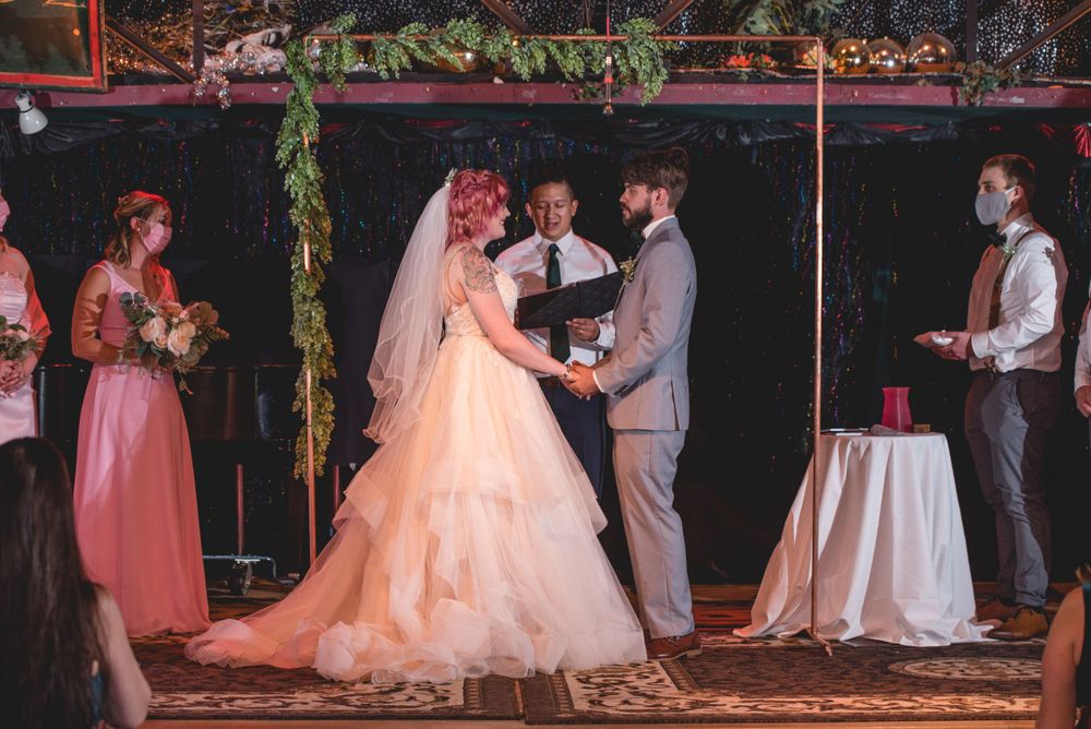 A wedding ceremony takes place in a dimly lit venue decorated with hanging greenery.