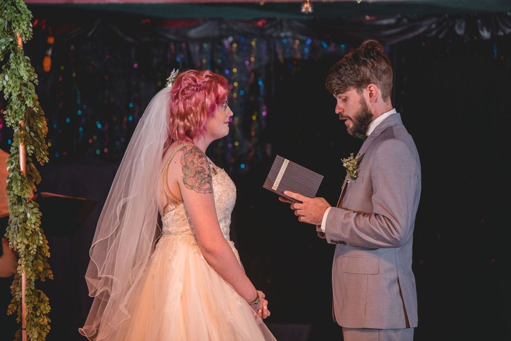 A couple exchanges vows during a wedding ceremony beneath twinkling lights and greenery.