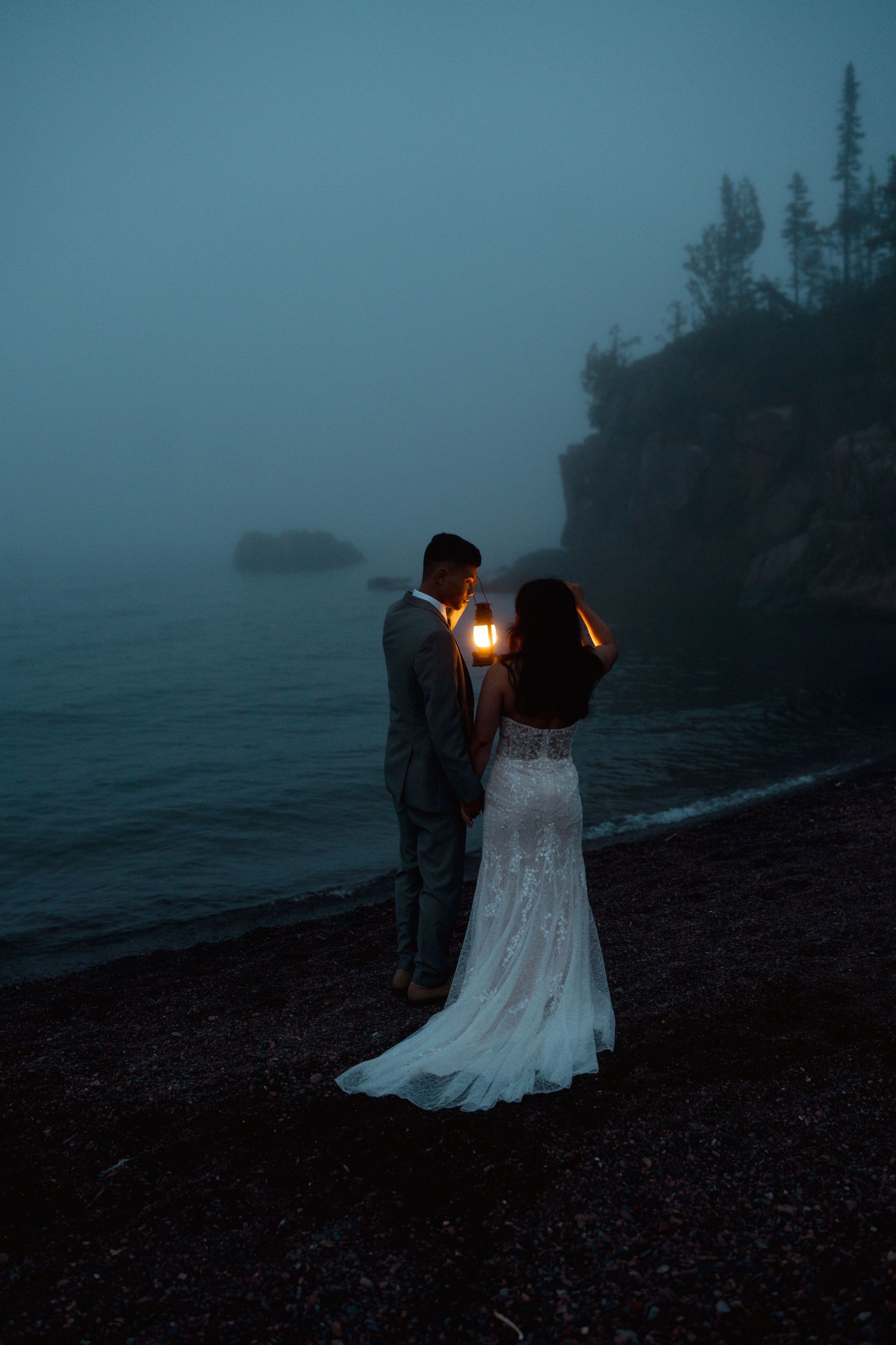 Bride and groom holding lantern on foggy beach at dusk, bride in lace wedding gown.