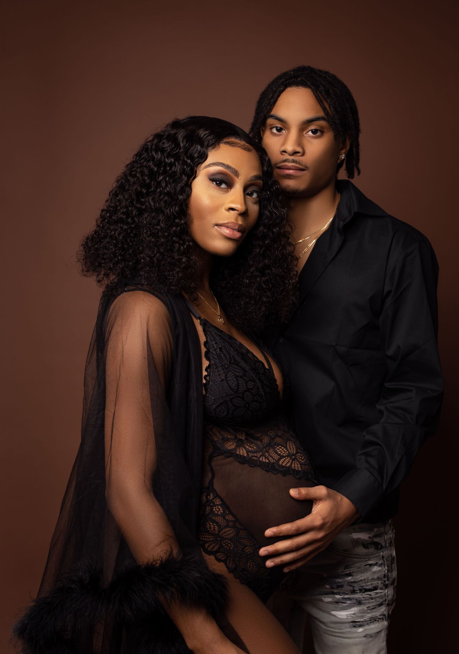 Elegant maternity portrait of couple in black attire against brown background.