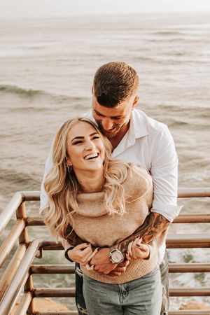 A joyful couple laughing together on wooden steps overlooking the ocean.