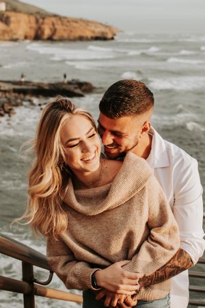 Joyful couple embracing on coastal overlook with crashing waves below.