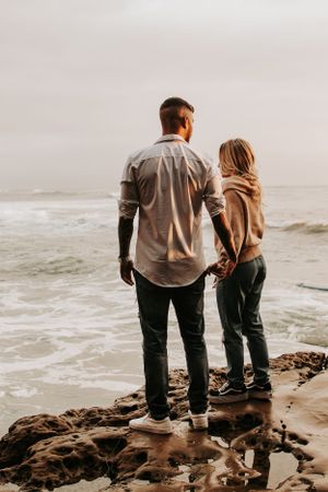 Couple standing on rocky shore overlooking ocean at sunset.