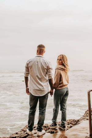 Couple stands together holding hands on rocky beach overlooking misty ocean waves.
