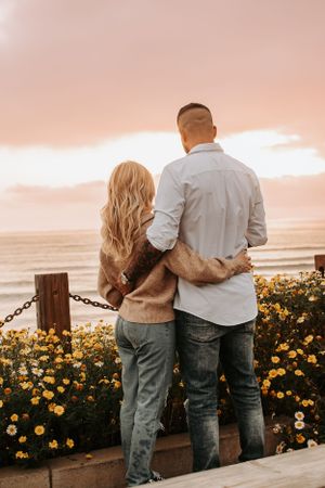 Couple watches sunset together among yellow wildflowers by ocean railing.