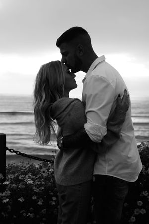 Black and white photo of couple embracing by ocean during sunset.