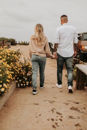 Couple walking together on sandy path beside blooming yellow flowers.