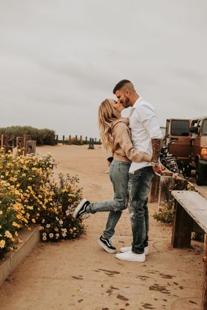 Romantic embrace on sandy path with yellow flowers and rustic wooden fence.