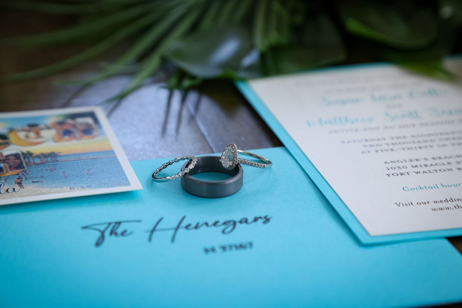 Wedding rings and invitation details displayed on a vibrant turquoise blue surface.