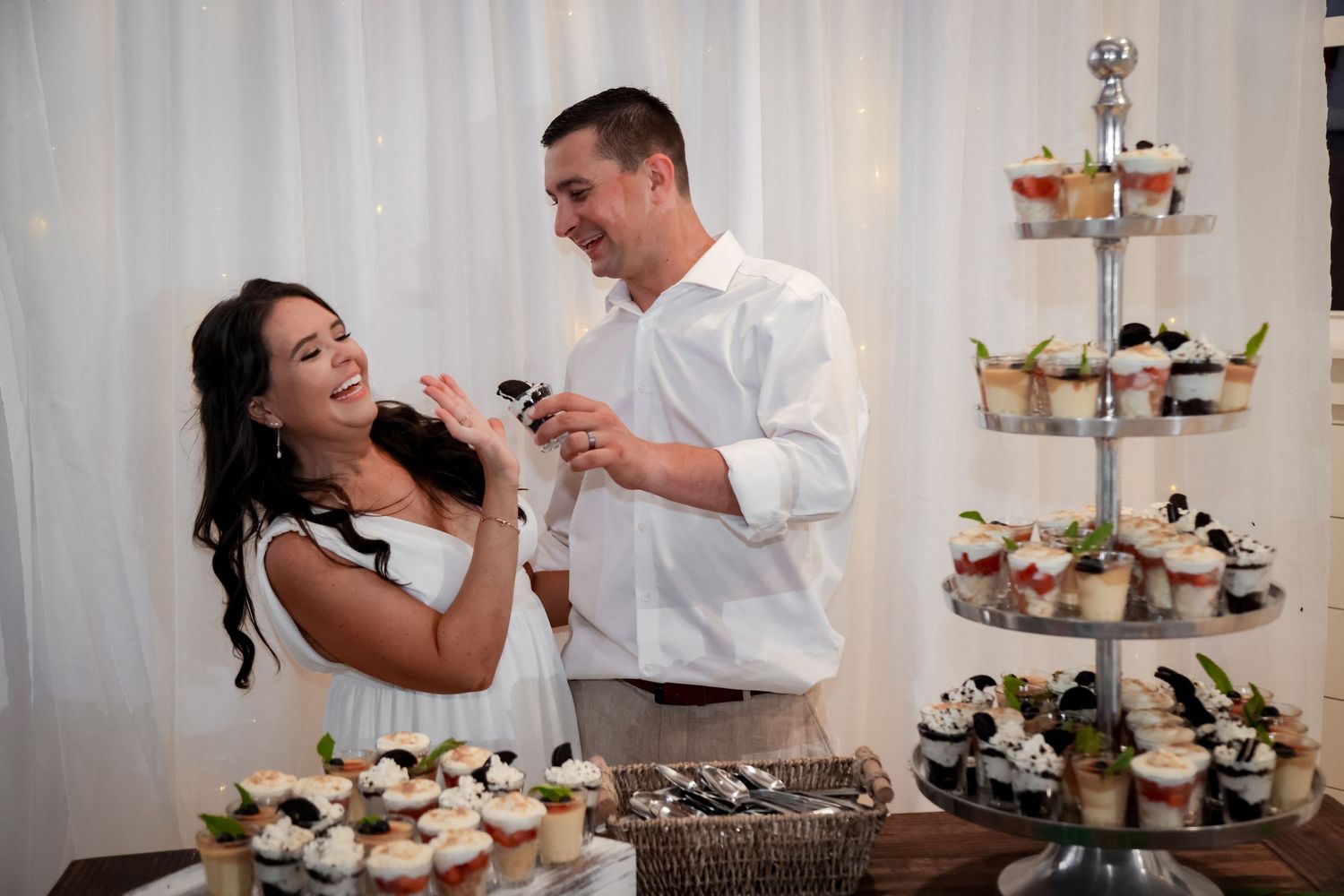 A joyful couple shares a laugh while cutting their wedding cake next to a tiered display of cupcakes.