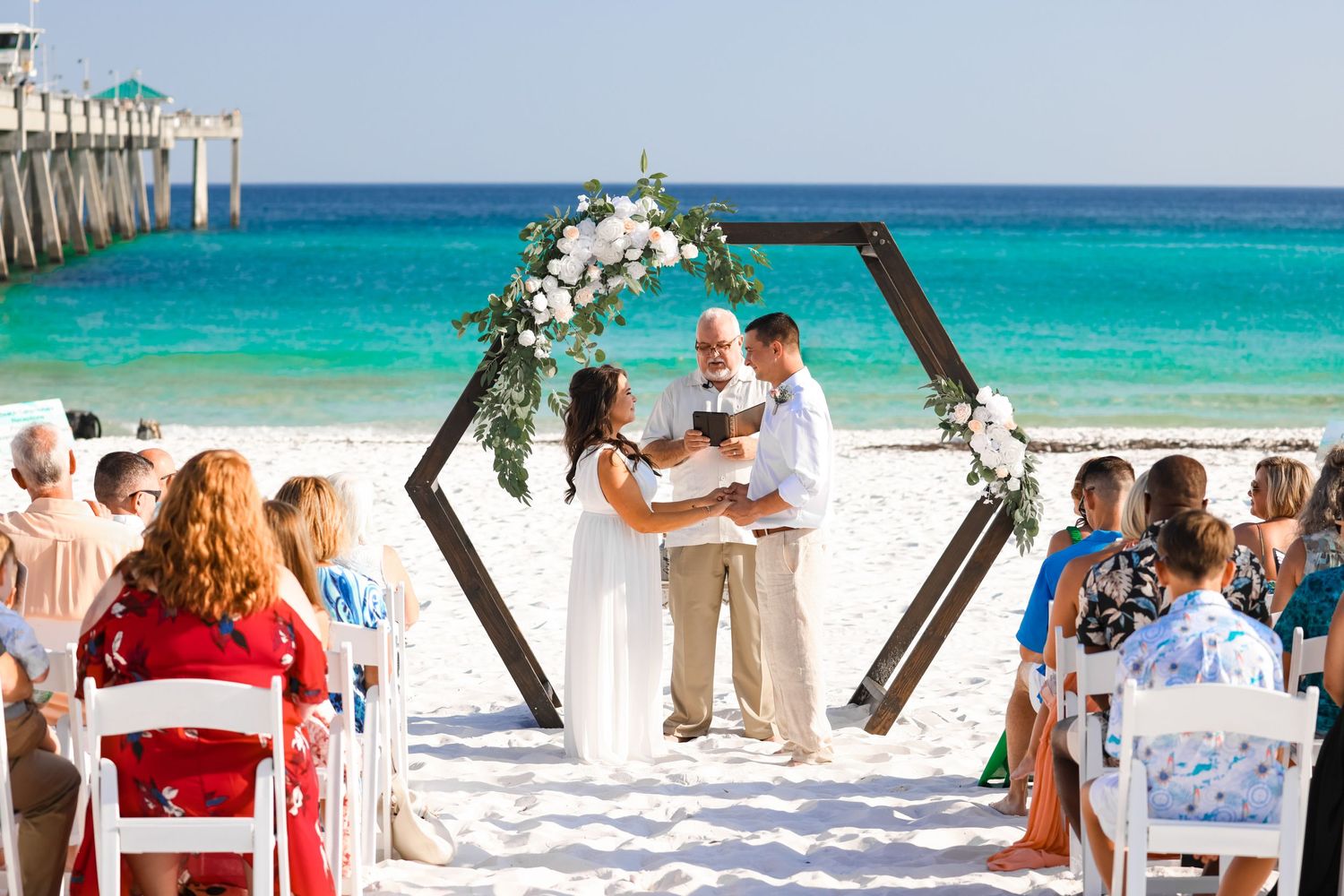 Beach wedding ceremony under a hexagonal arch with turquoise ocean and white sand backdrop.