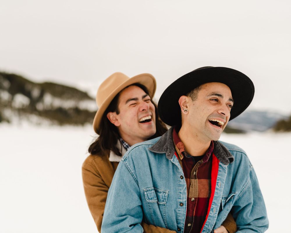 Two people wearing hats laughing together in a snowy winter landscape.