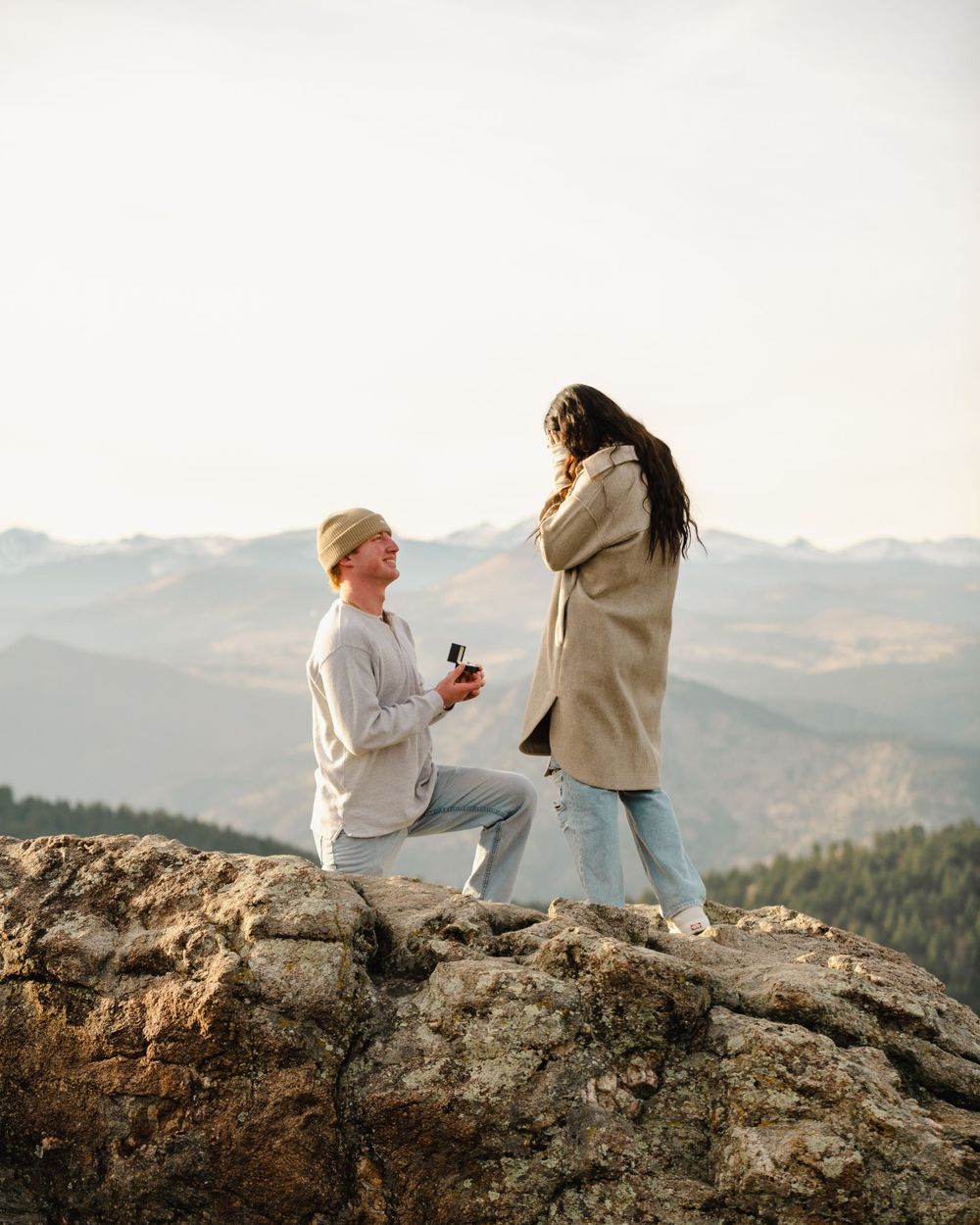 Surprise proposal moment on a mountaintop during sunset with scenic mountain views.