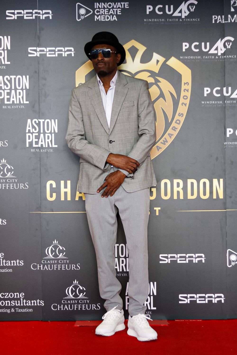 A figure in a gray suit and sunglasses poses on a red carpet with event sponsor logos in the background.