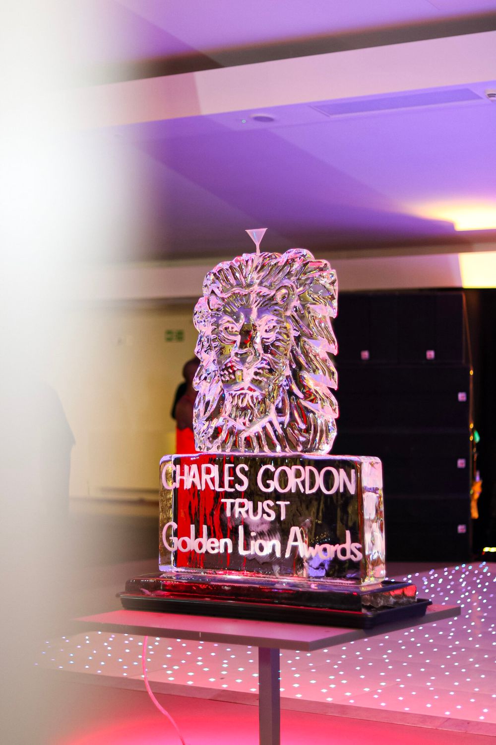 An illuminated ice sculpture of a lion head with Charles Gordon Trust text displayed on a red base.