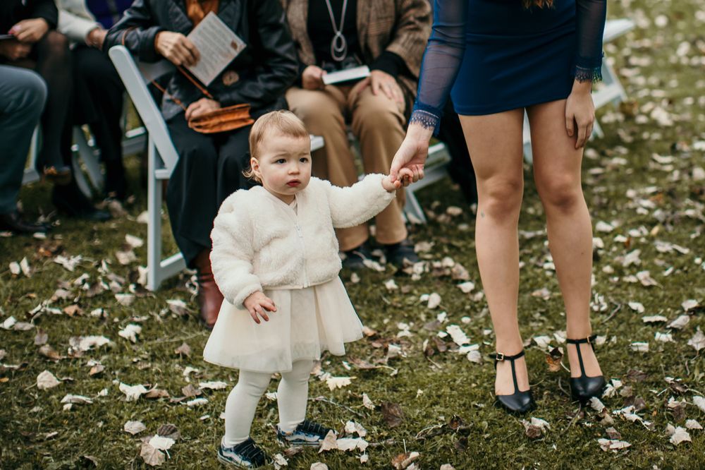 A small child in a white dress holds hands with an adult during an outdoor wedding.
