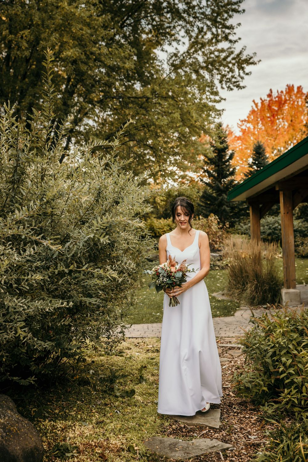 A white wedding dress and bouquet stand out against autumn foliage in a garden setting with warm golden light.