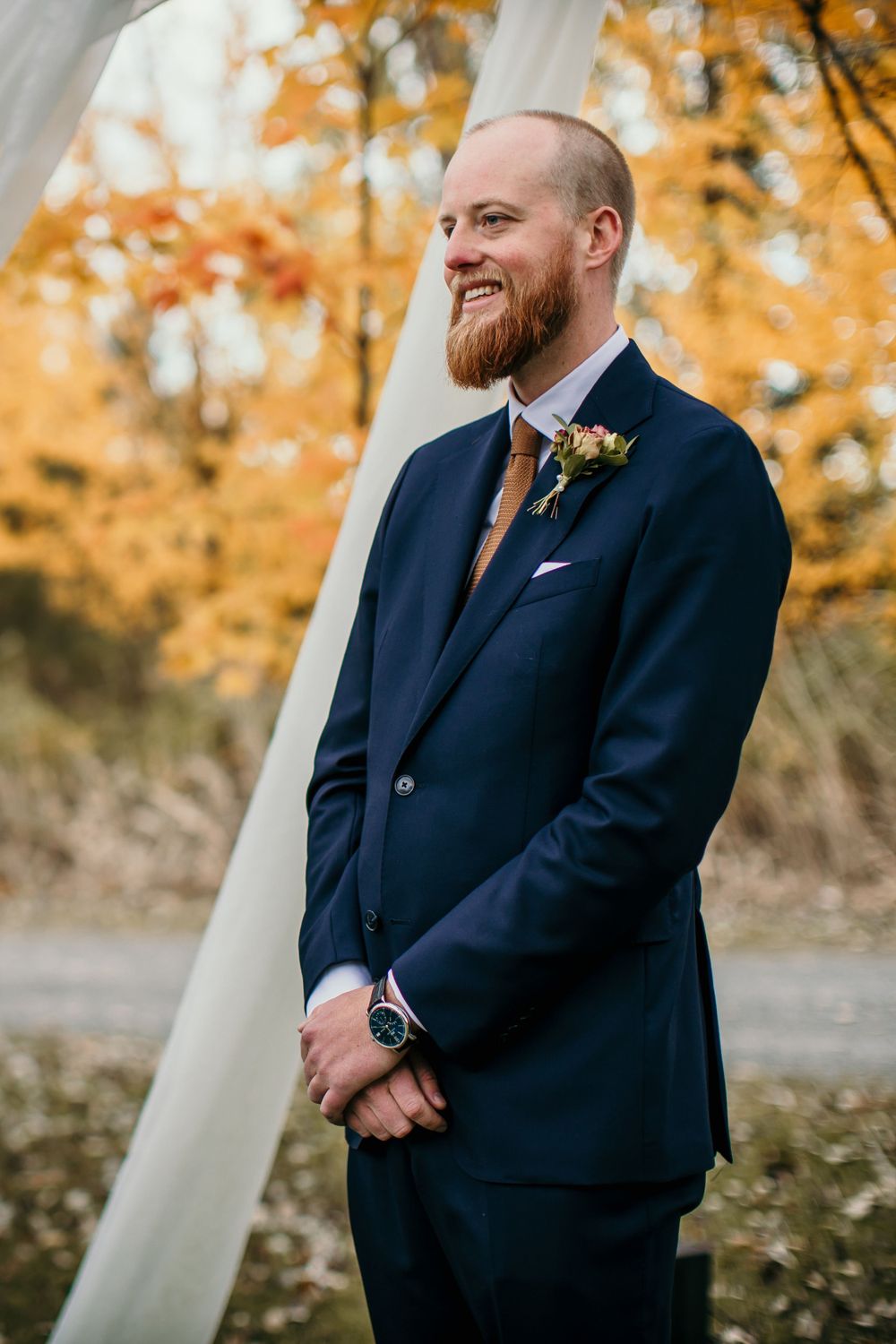 A groom in a navy suit smiles during an autumn wedding ceremony.
