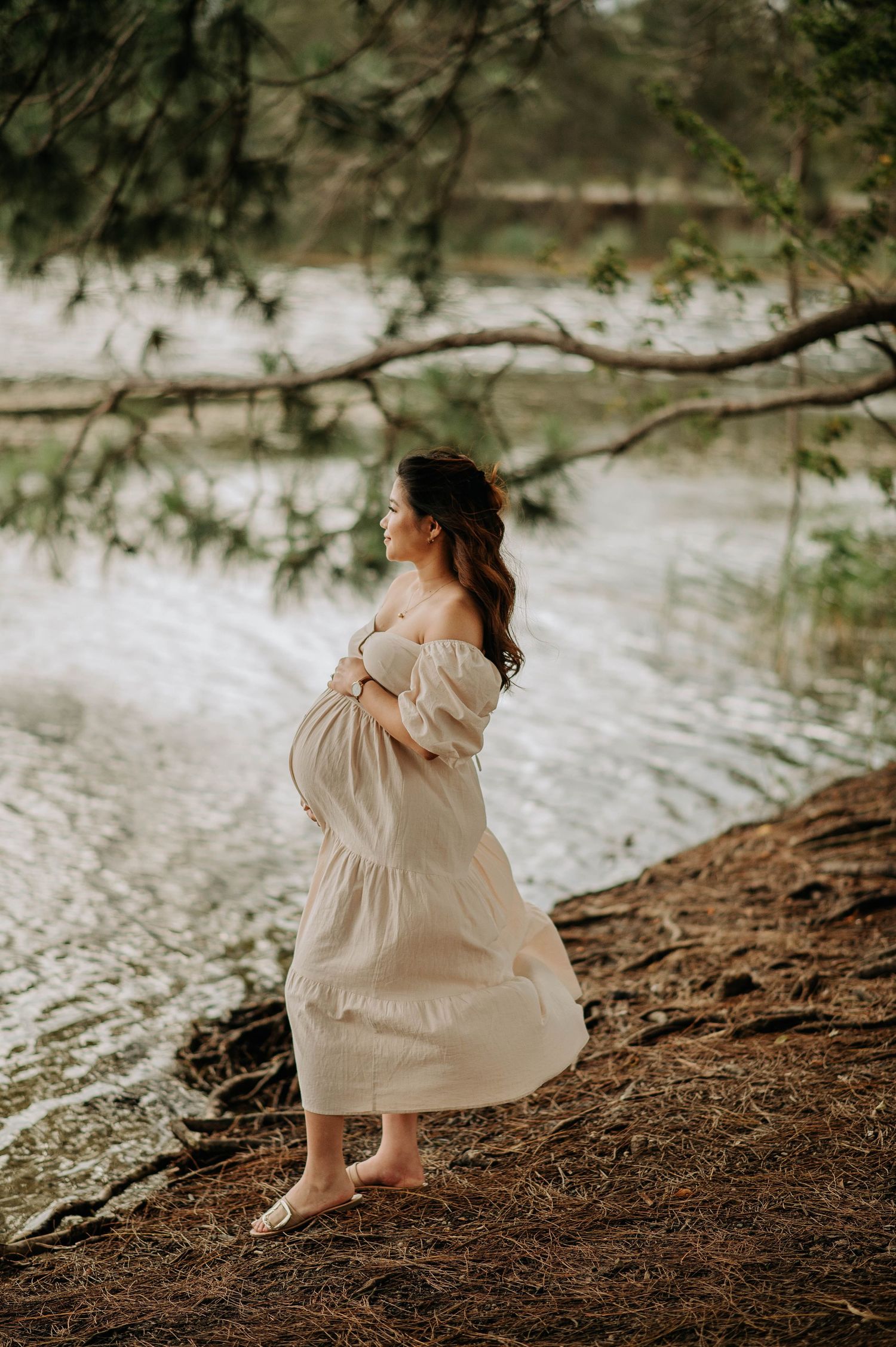 Pregnant woman in flowing dress stands barefoot by waterside during golden hour.