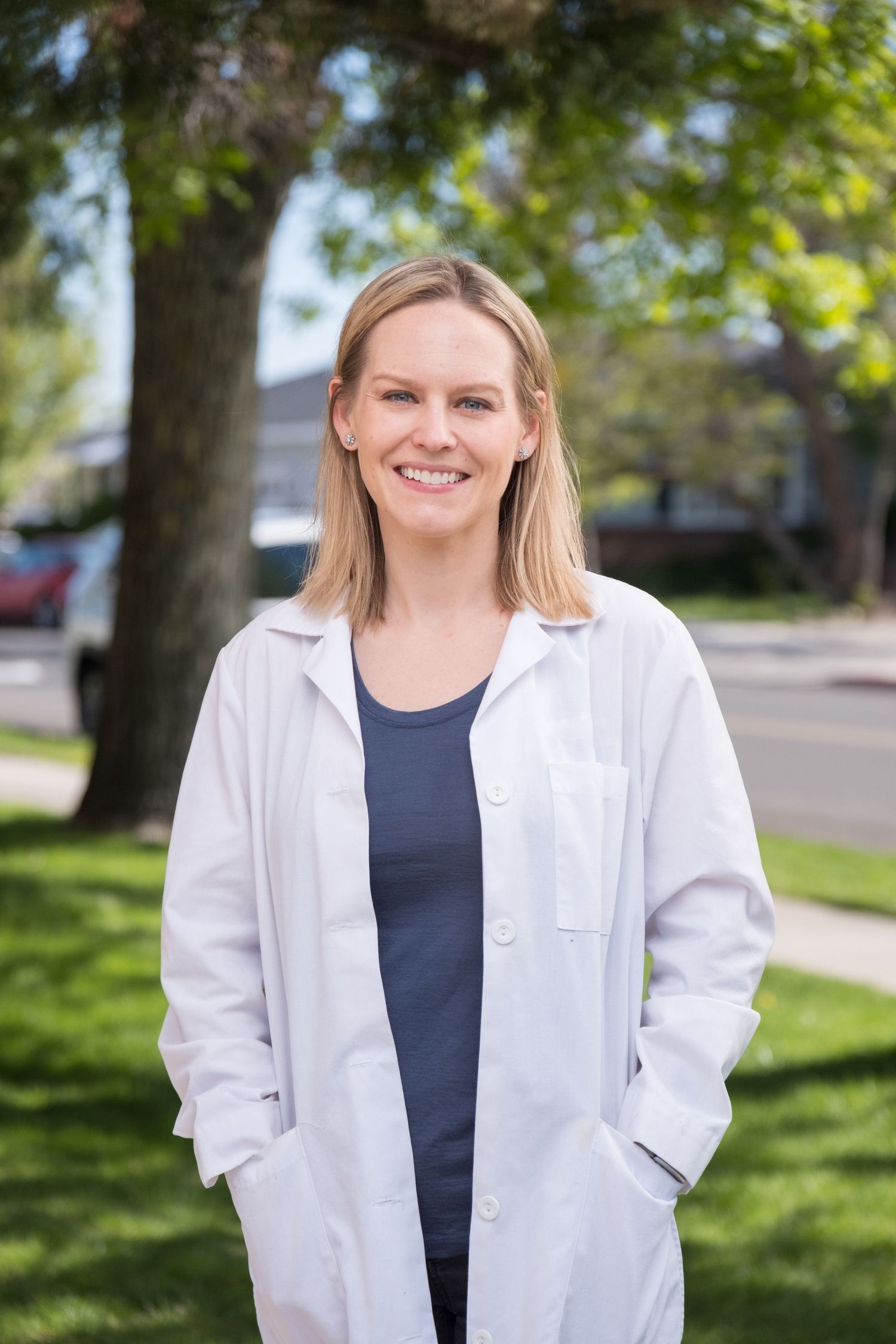 Medical professional in white lab coat and navy shirt stands outdoors on sunny day with trees in background.