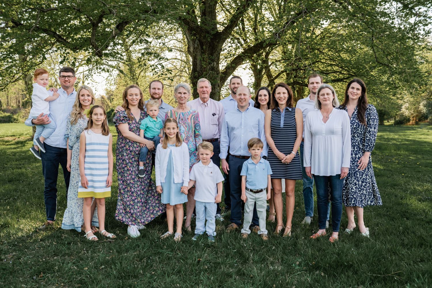 Large family group portrait with multiple generations posed outdoors under trees.