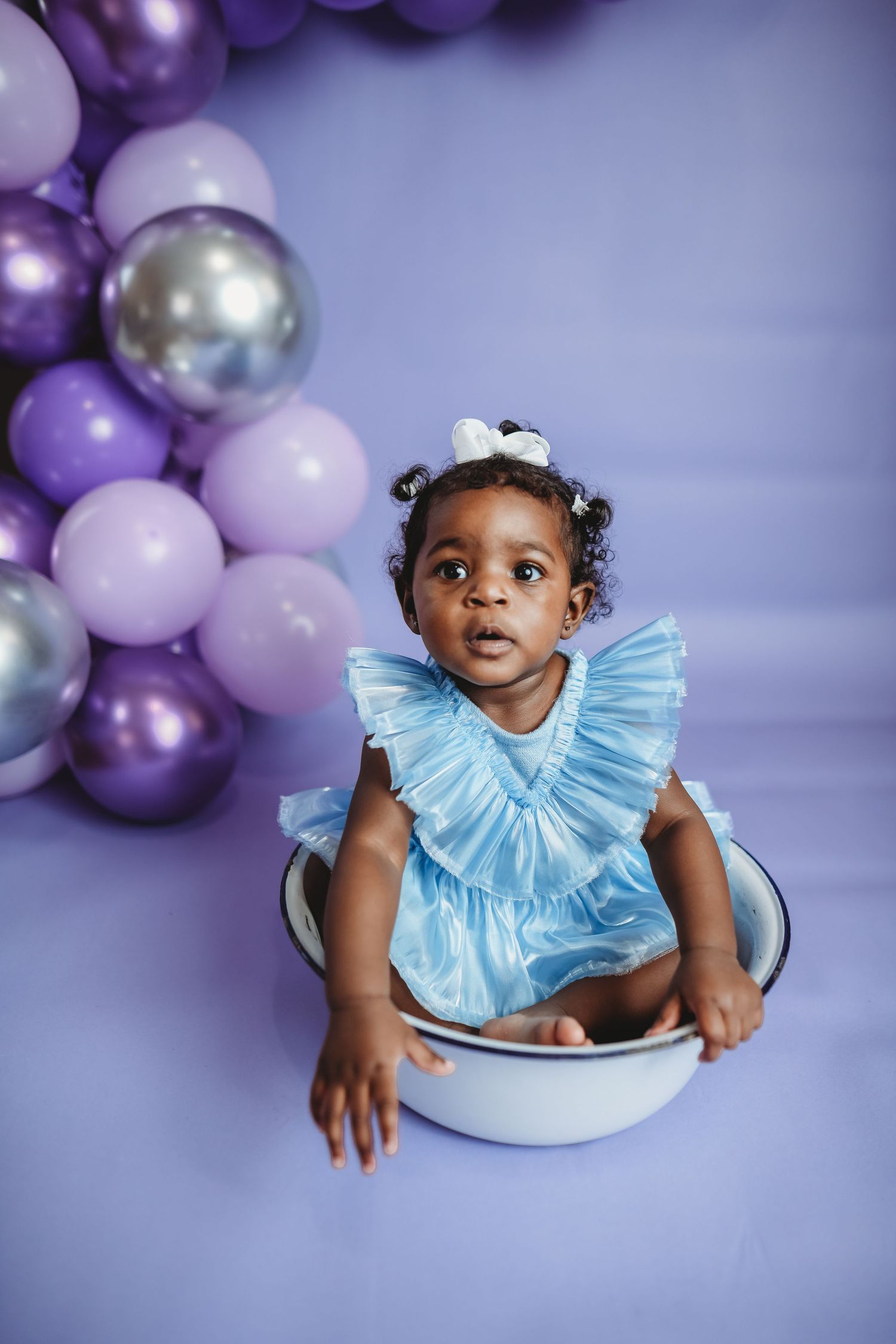 Baby in light blue ruffle dress sitting in basin with purple and silver balloon arch backdrop.