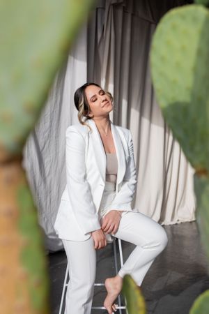 Woman in white suit poses elegantly near indoor plants.