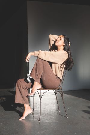 Woman in beige sweater and brown pants posing on chair in dramatic lighting.