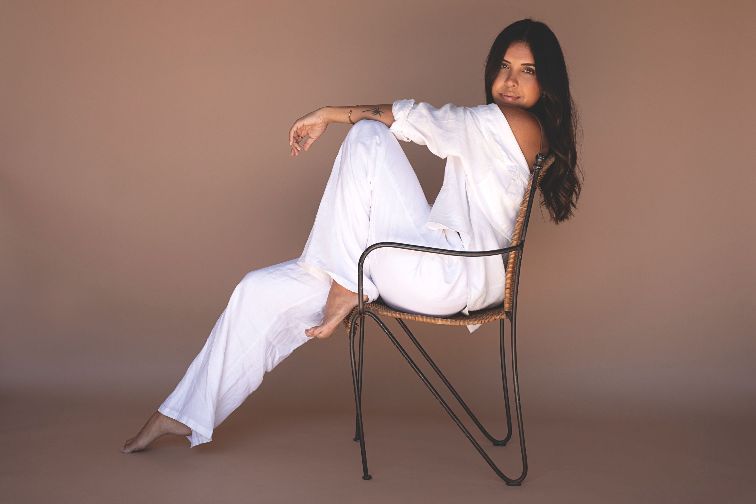 Woman in white outfit lounging gracefully on metal chair against pink backdrop.