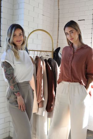 Two people in casual attire standing by a clothing rack with neutral toned garments.