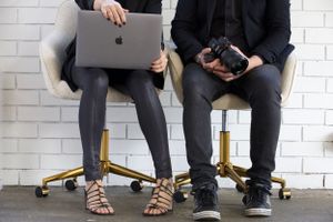 Two people sitting with laptop and camera against white brick wall.