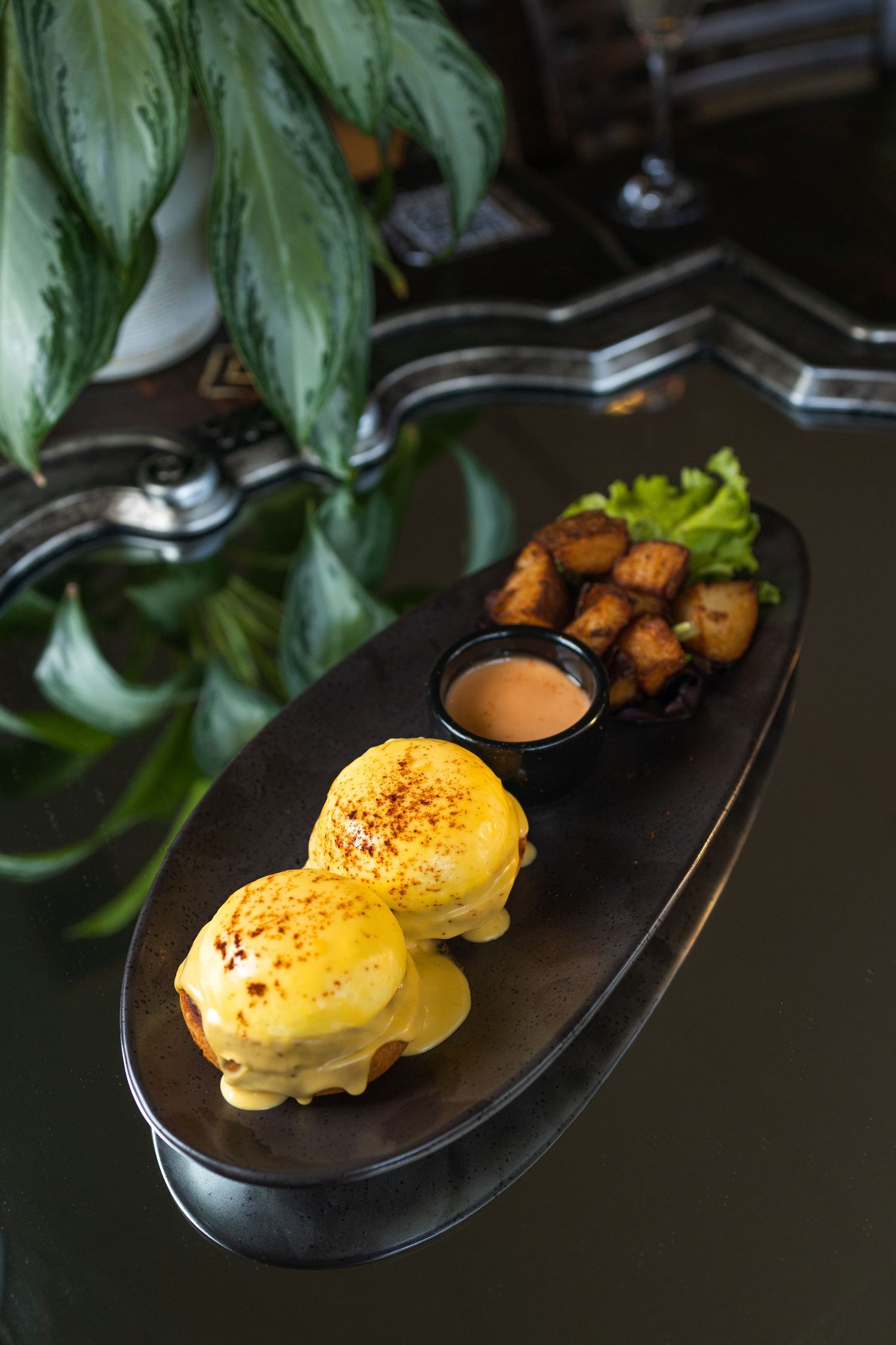 Eggs Benedict with hollandaise sauce and roasted potatoes served on a black oval plate with fresh herbs.