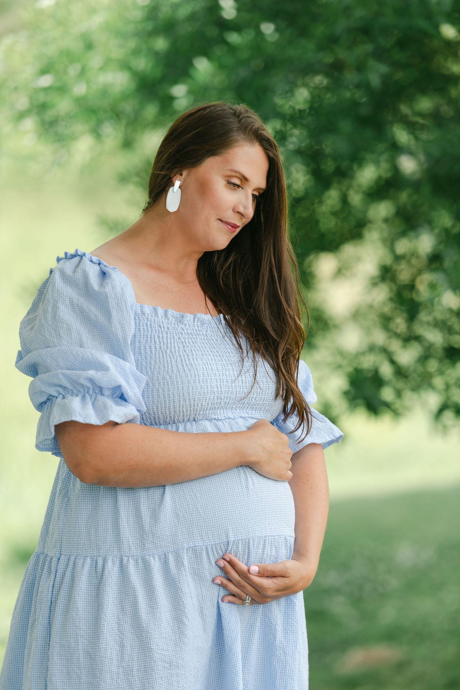 Pregnant woman in blue smocked dress holding baby bump while standing in lush green park setting.