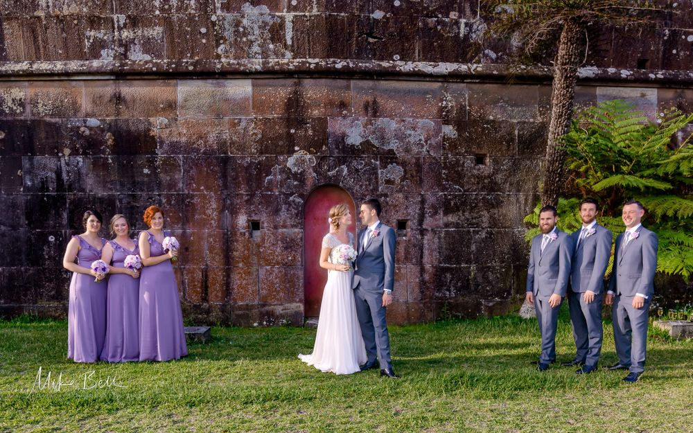 A wedding party poses against a rustic stone wall with bridesmaids in purple dresses.
