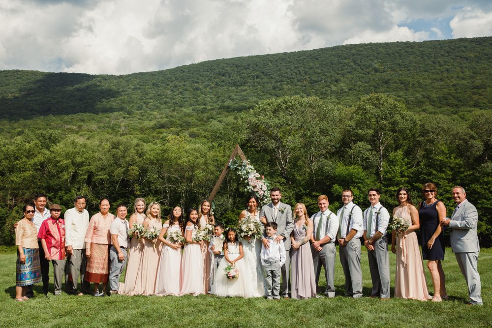 Large wedding party group photo outdoors with mountain backdrop and wooden triangle arch.