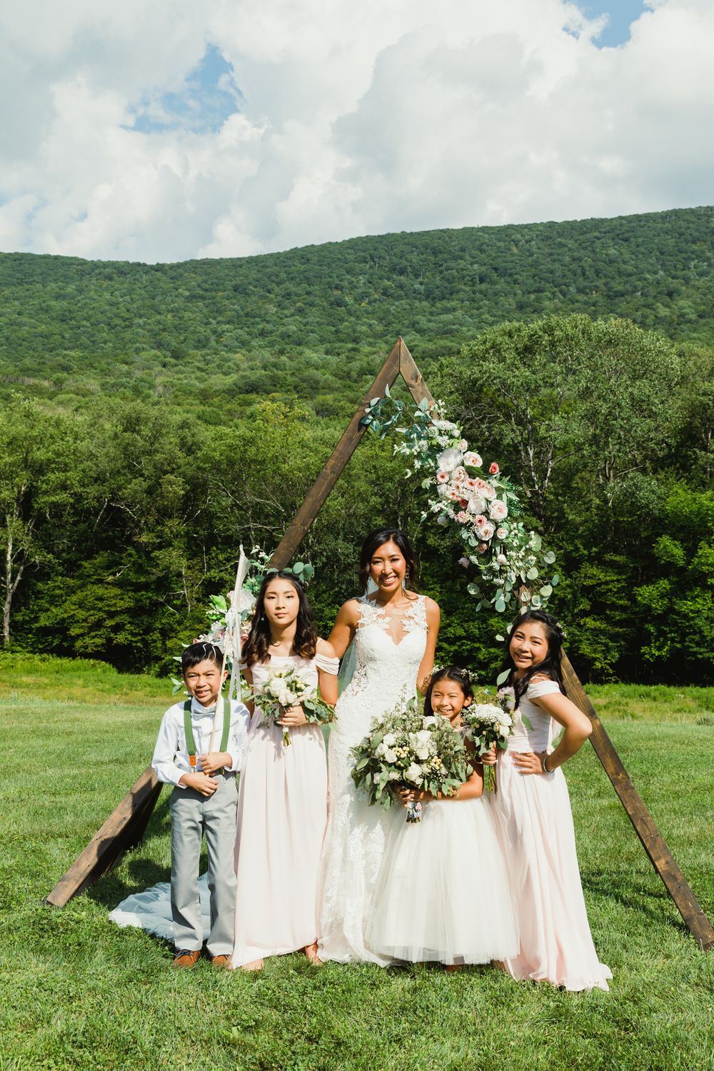 Wedding party members posing with floral-decorated wooden triangle arch against mountain landscape.